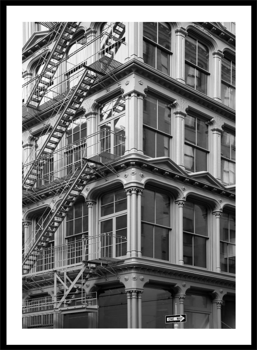 Black and white architectural photograph of a corner building facade with ornate windows and external fire escapes. Urban Geometry by Shot by Clint black-and-white architectural photography print featuring fire escapes, tall windows, ornate columns, and layered city detail.