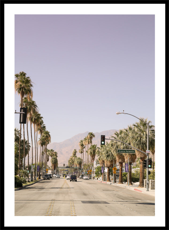 A framed art print featuring a scenic photograph of a palm-lined street in Palm Springs with mountains in the background. Palm-Lined Avenue by Shot by Clint travel photography print featuring a California road, tall palm trees, desert mountains, and soft sun-faded tones.