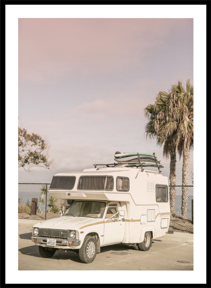 A vintage white camper truck parked outdoors near palm trees and a fence, with a surfboard strapped to its roof rack. Vintage Camper Parked by Shot by Clint travel photography print featuring a retro camper, surfboards, palm trees, and sun-faded coastal tones.