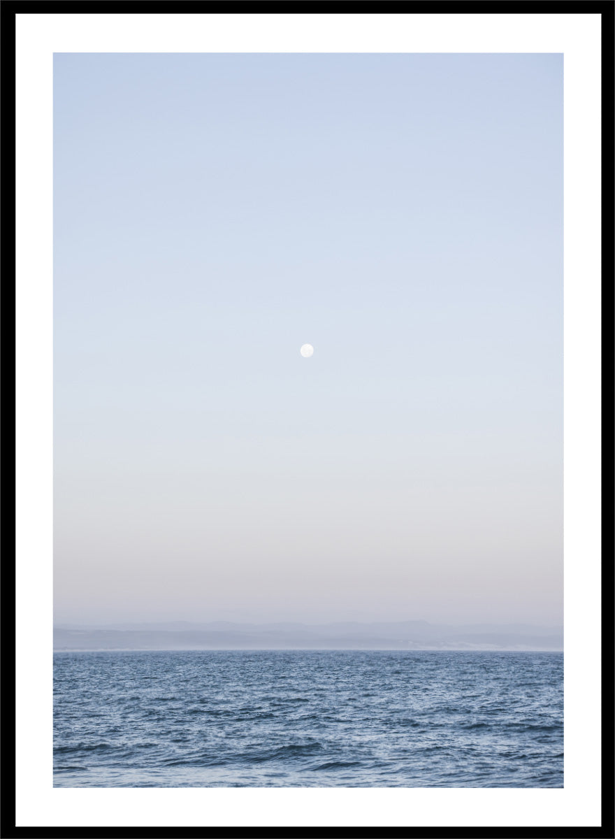 A minimalist framed art print depicting a calm blue ocean under a pale sky with a small visible moon. Moonlit Horizon by Shot by Clint coastal photography print featuring a pale moon, distant shoreline, open blue water, and soft lavender evening tones.