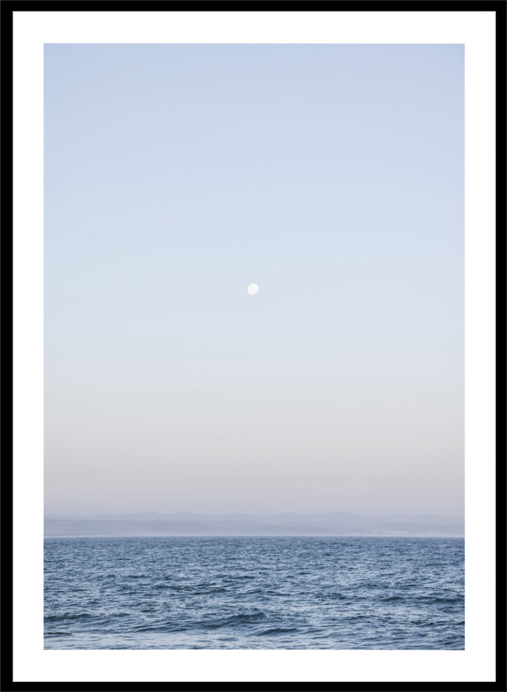 A minimalist framed art print depicting a calm blue ocean under a pale sky with a small visible moon. Moonlit Horizon by Shot by Clint coastal photography print featuring a pale moon, distant shoreline, open blue water, and soft lavender evening tones.
