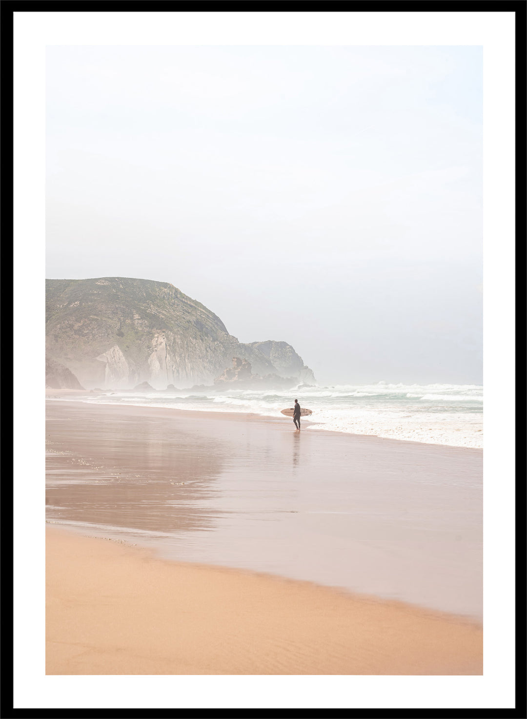 Person walking along a beach with mountains in the background. Misty Morning Surfer by Henrike Schenk captures a surfer walking along a misty beach with ocean waves, coastal scenery, and photography wall art.