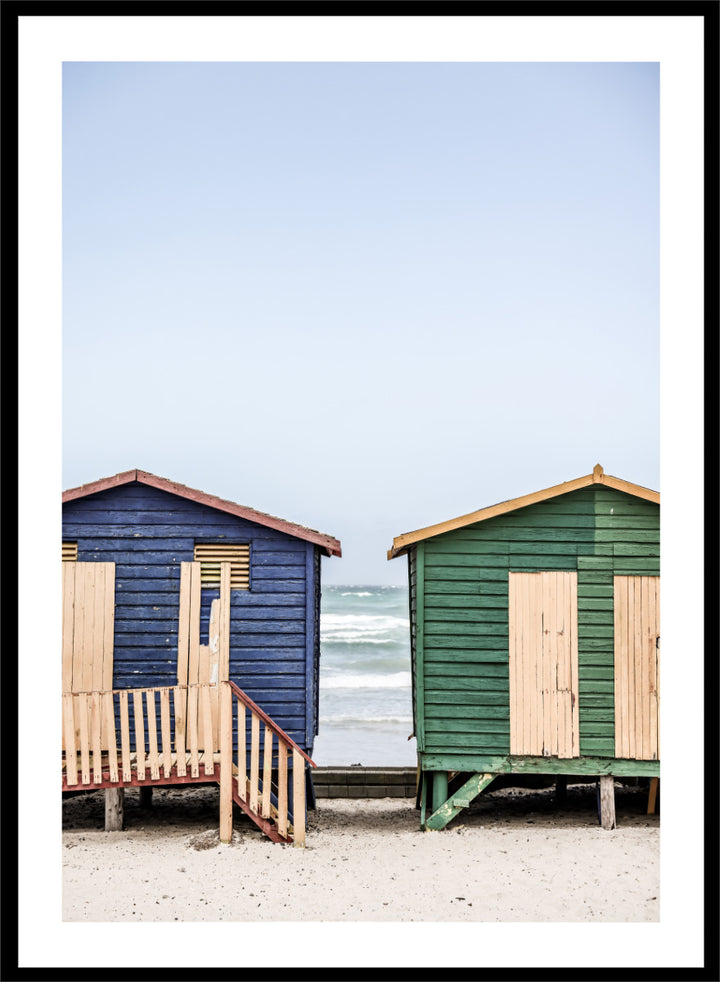 A framed photographic print of two wooden beach huts on a sandy beach under a pale blue sky, with one hut in dark blue and the other in green, both with light wood shutters and doors. Coastal Huts Harmony by Shot by Clint coastal photography print featuring blue and green beach huts, sandy ground, weathered wood, and ocean views.