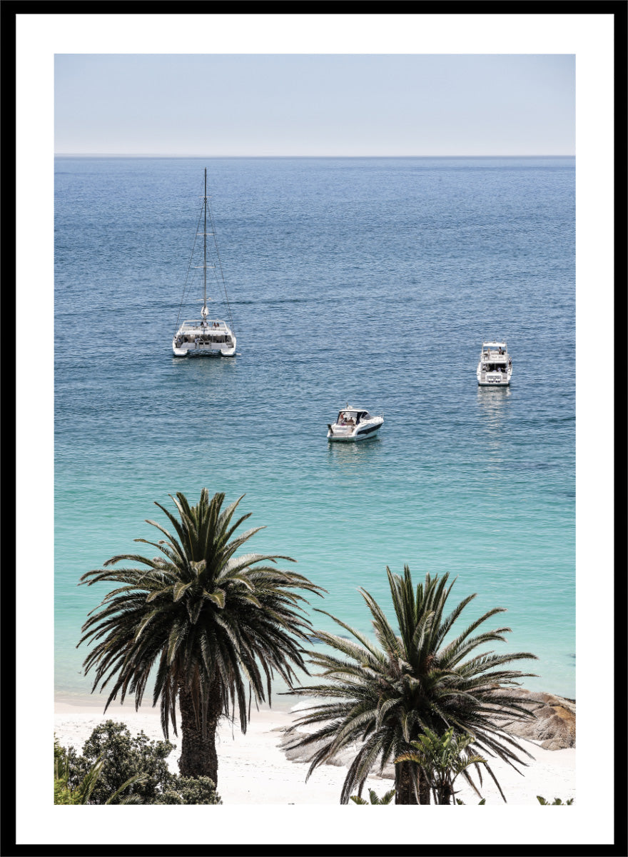 A framed coastal photograph showing a calm blue ocean with three boats, turquoise water near a white sandy shore, and two palm trees in the foreground. Tropical Serenity by Shot by Clint coastal photography print featuring turquoise water, sailboats, palm trees, and a bright tropical shoreline.