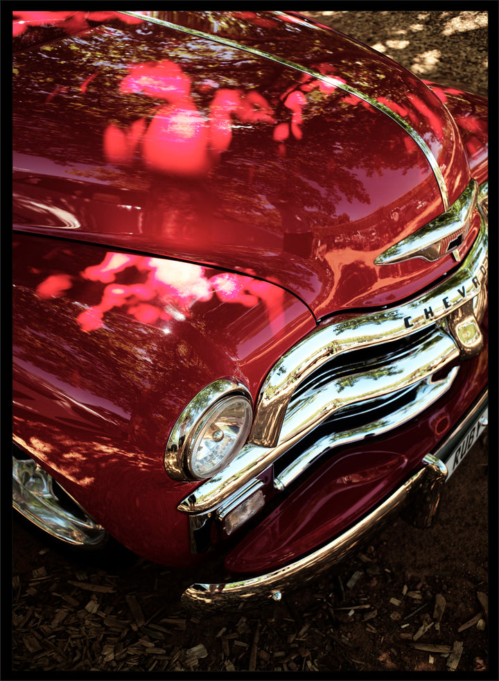 Close-up of a vintage red Chevrolet car body with chrome grille and reflections of trees on the glossy paint, photographed outdoors on mulch. Vintage Red Car by Shot by Clint automotive photography print featuring a classic Chevrolet, deep red paint, polished chrome, and sculptural vintage details.