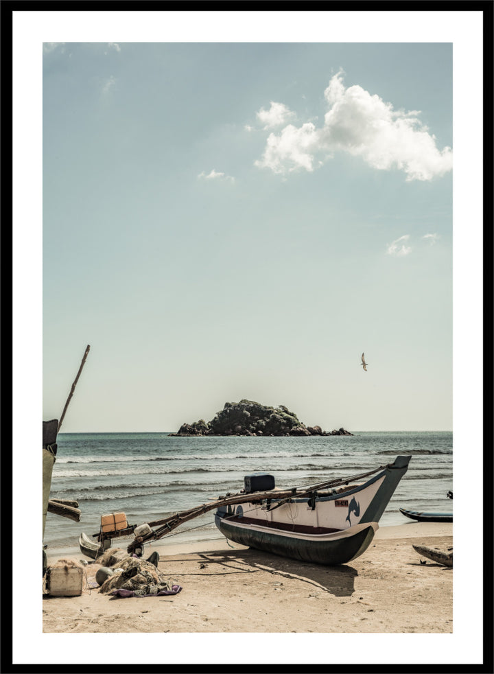A framed photographic print of a coastal beach scene with a small boat on the sandy shore, the sea and distant rocky island, and a pale blue sky with clouds. Seaside Serenity by Shot by Clint coastal photography print featuring a fishing boat, sandy shore, ocean waves, distant island, and weathered seaside details.