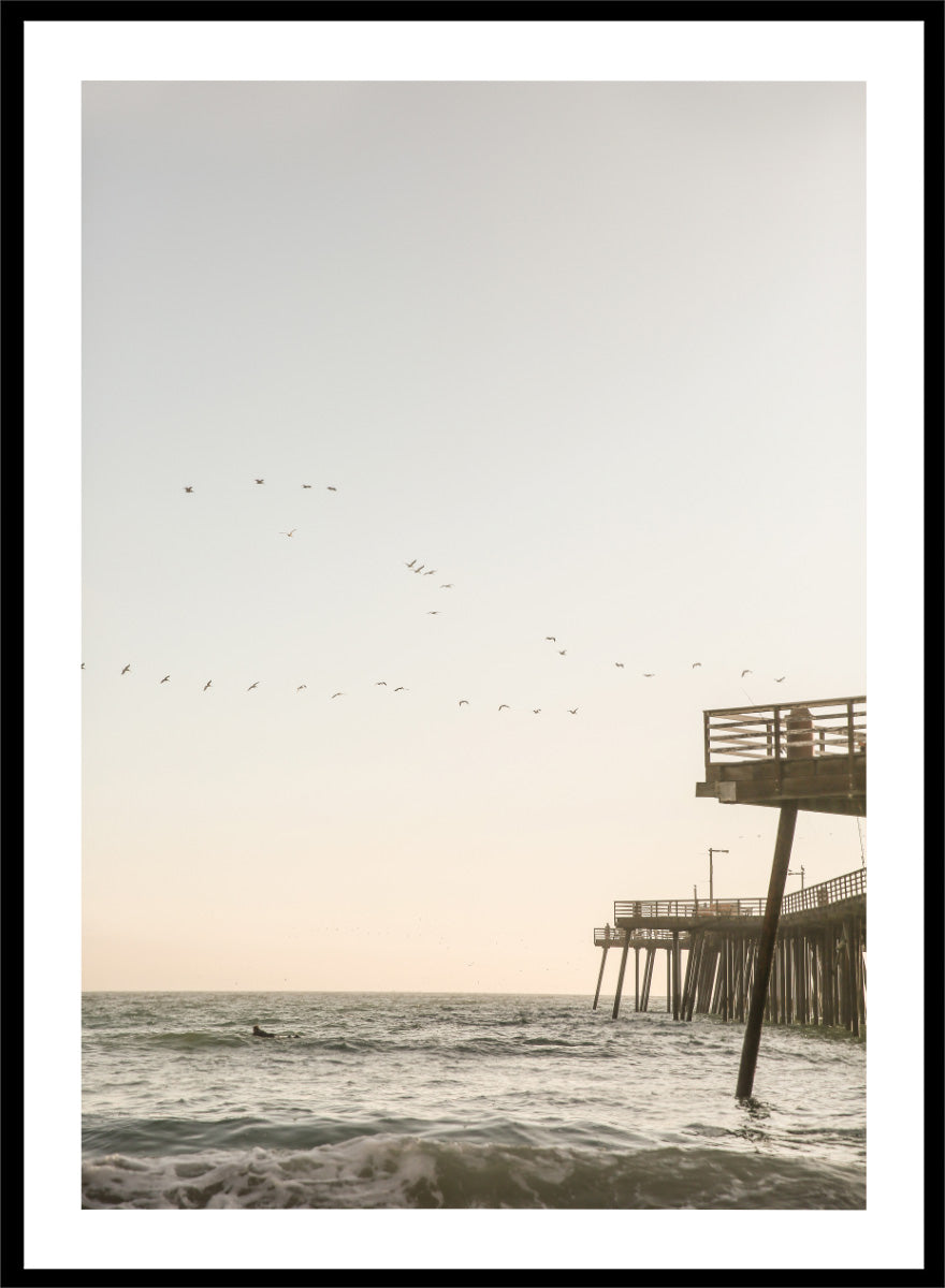 A framed photographic art print depicting a pier extending over a calm ocean under a pale sky, with birds flying across the scene. Seaside Serenity by Shot by Clint coastal photography print featuring ocean waves, a weathered pier, flying birds, and soft sunlit tones.