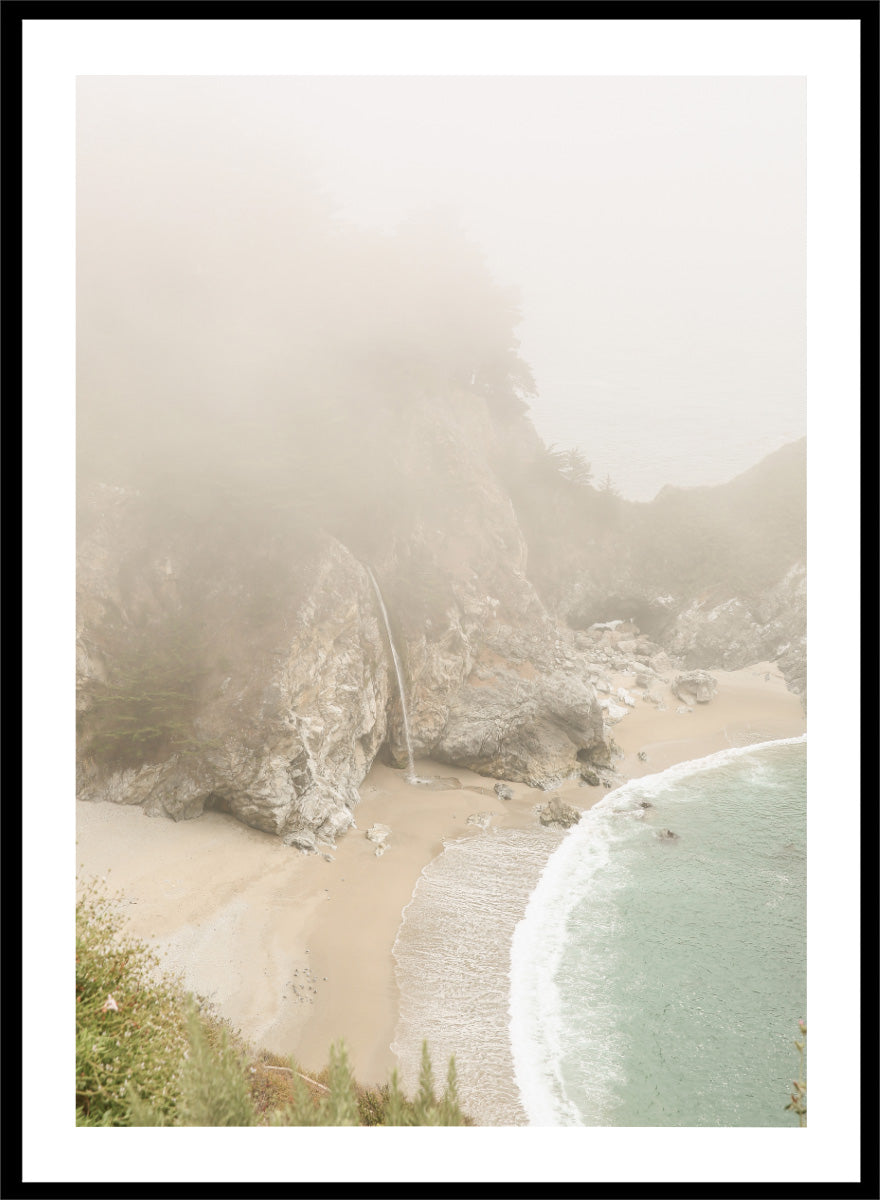 A framed photographic print of a misty coastal beach scene with a small waterfall, sandy shore, and pale blue-green ocean waves. Misty Coastal Haven by Shot by Clint coastal photography print featuring a secluded cove, rocky cliffs, pale turquoise water, and a soft veil of mist.