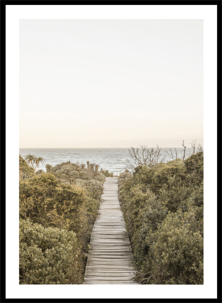 A framed photographic art print depicting a wooden boardwalk leading from dense green beach vegetation to the ocean under a pale sky. Pathway to Tranquility by Shot by Clint coastal art print featuring a wooden boardwalk, wild greenery, distant palms, and a soft ocean horizon.