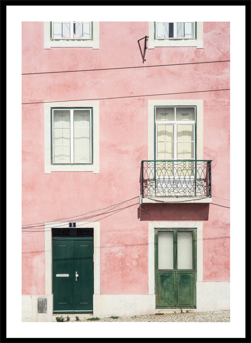 A framed art print featuring a photograph of a pink building facade with green doors, white-framed windows, and a small black metal balcony. Pink Façade Harmony by Shot by Clint architectural photography print featuring a pink exterior, green doors, white windows, and an iron balcony.