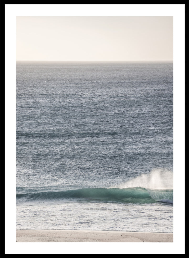 A framed photographic art print of an ocean scene, featuring a gentle wave breaking on a sandy beach under a pale sky. Tranquil Ocean Scene by Shot by Clint ocean photography print with a rolling wave, soft sea spray, and airy blue coastal tones.