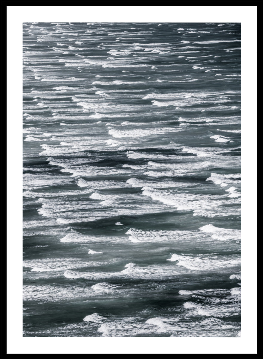 A framed black and white photographic print depicting a close-up view of ocean waves with white foam crests against dark water. Waves in Rhythm by Shot by Clint ocean photography print featuring layered blue-grey waves, white foam, and powerful natural movement across the sea.