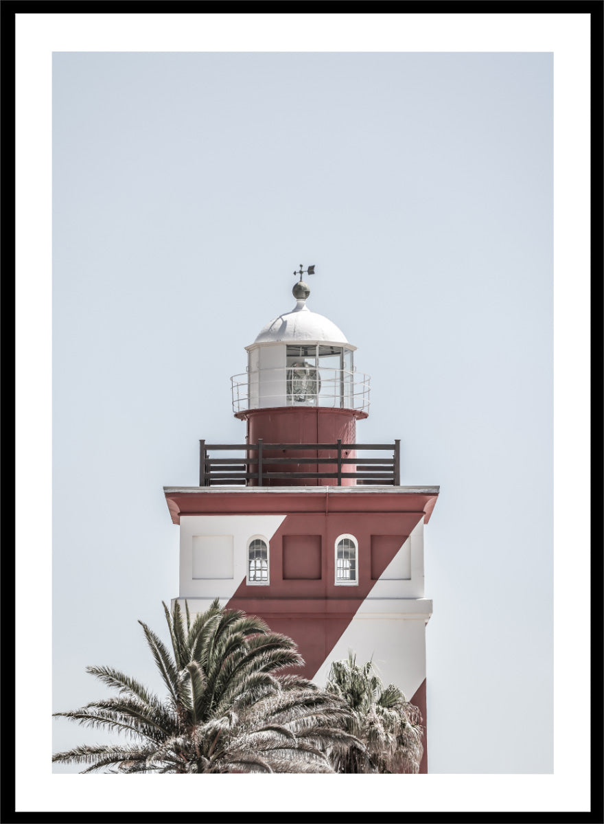 A framed art print featuring a red and white lighthouse with palm trees at the bottom, set against a pale sky background. Beacon of Solitude by Shot by Clint lighthouse photography print featuring red-and-white architecture, palm trees, arched windows, and a pale coastal sky.