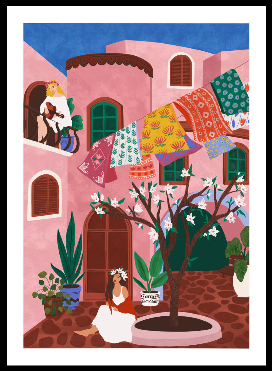 A framed art print featuring a colorful Mediterranean-style illustration of a pink courtyard with a woman sitting under a flowering tree, laundry hanging in the background, and a blue sky above. Pink Village Courtyard by Ceyda Alasar art print featuring pink architecture, patterned rugs, flowering trees, potted plants, and relaxed courtyard life.