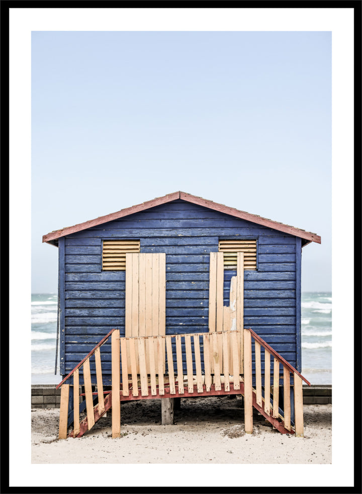 A framed photographic print of a blue wooden beach lifeguard hut on a sandy shore with the ocean and pale sky in the background. Beachside Retreat by Shot by Clint features a weathered blue beach hut and soft ocean backdrop in a calm coastal photography print.