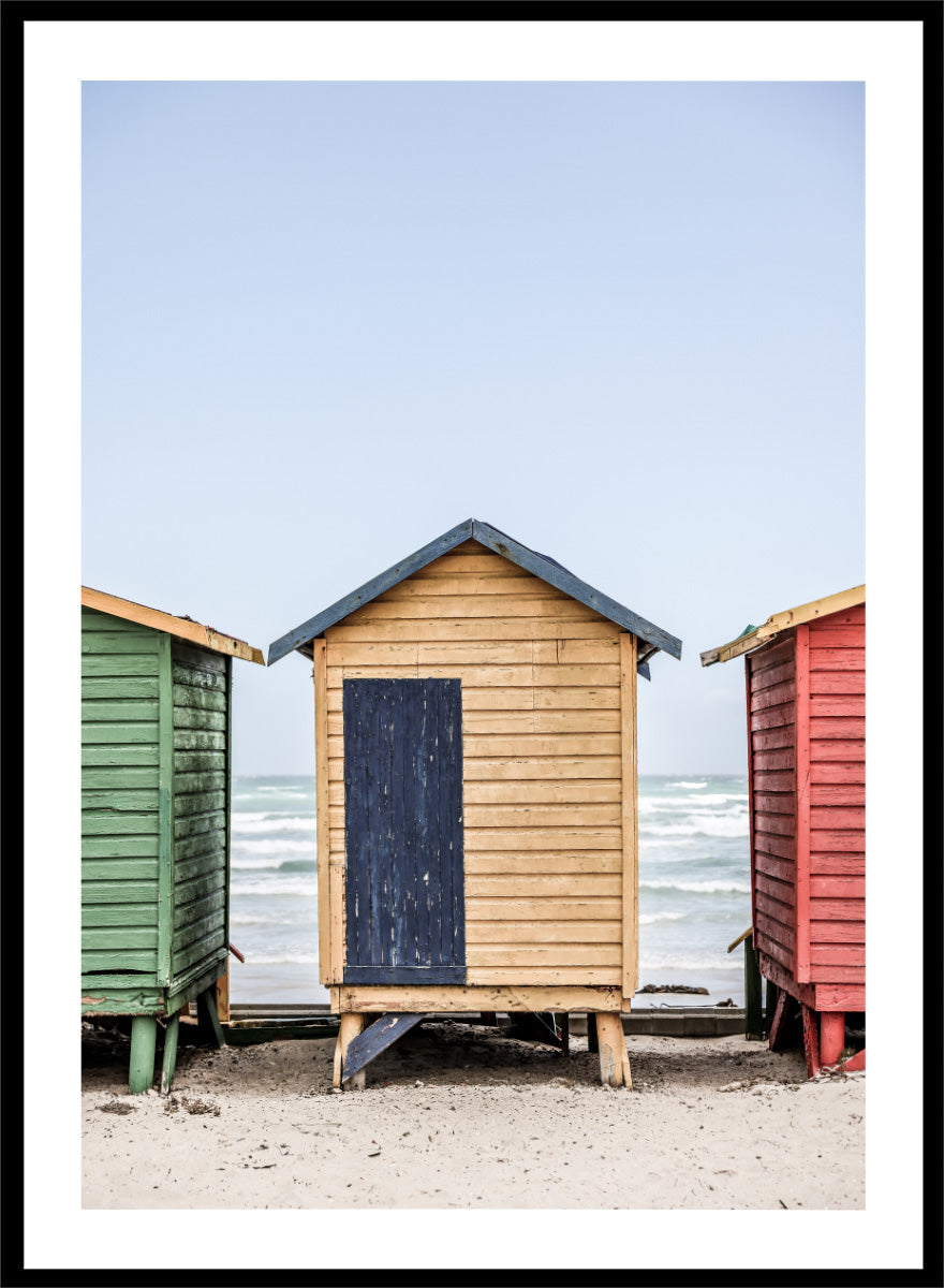 A framed photographic print of three colorful wooden beach huts on a sandy beach, with the central hut in natural wood and a dark blue door, flanked by green and red huts under a pale blue sky. Colorful Beach Huts by Shot by Clint coastal photography print featuring green, natural wood, and red huts with weathered texture and ocean views.
