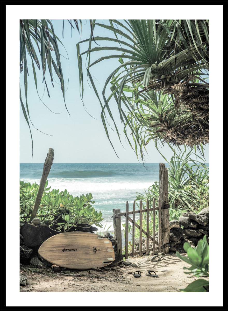 A framed beach scene art print featuring a wooden surfboard leaning against a rustic gate, with palm leaves framing a view of the ocean under a light blue sky. Coastal Serenity by Shot by Clint coastal art print featuring a surfboard, tropical plants, rustic gate, sandy path, and ocean waves.