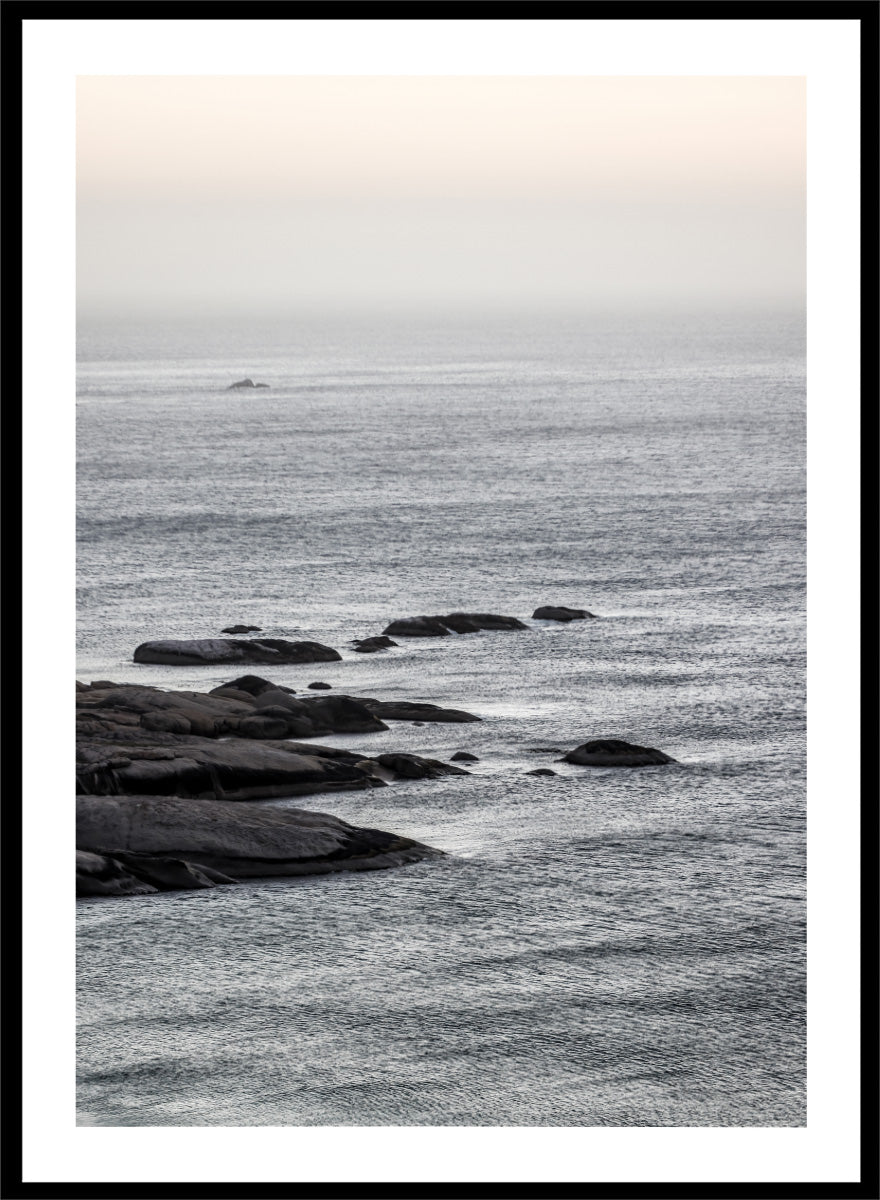 A framed black and white coastal landscape photograph featuring a calm sea, distant rocks, and a pale sky. Silver Sands by Shot by Clint coastal photography print featuring silver-blue water, dark shoreline rocks, and a soft misty horizon.