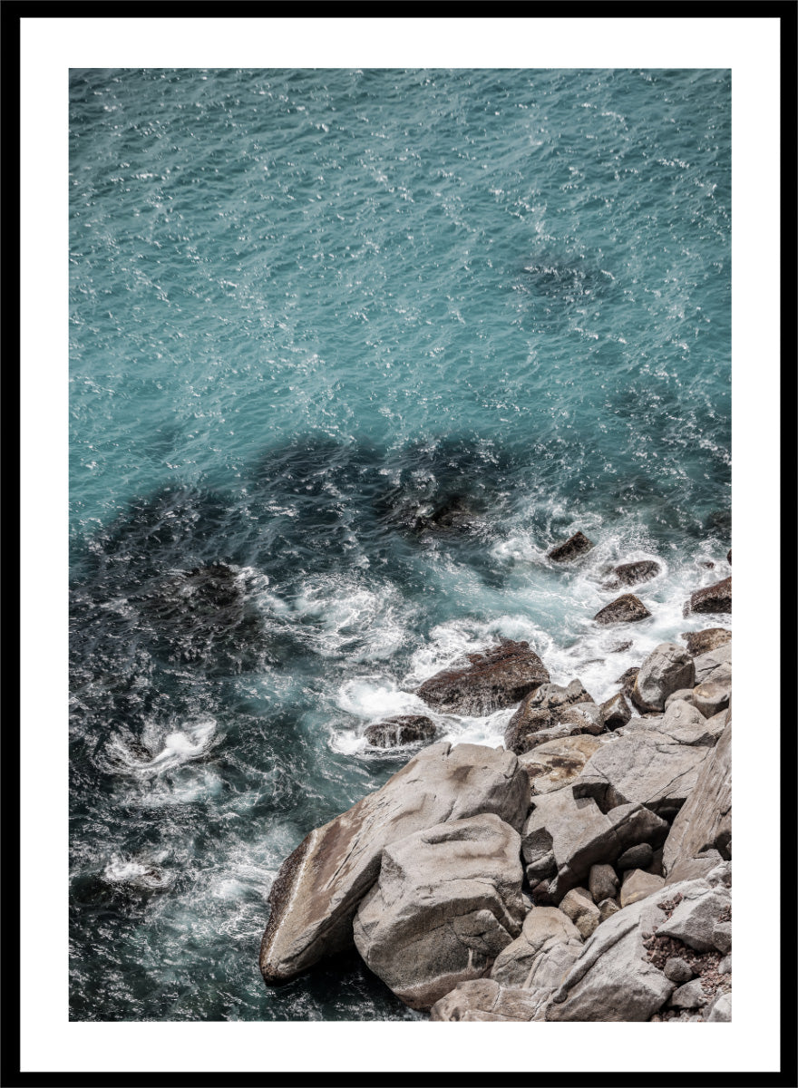 A framed photographic art print depicting a top-down view of turquoise ocean water washing over dark rocks and a rocky shoreline. Ocean’s Edge Symphony by Shot by Clint aerial ocean photography print featuring turquoise water, white surf, coastal texture, and soft seafoam tones.