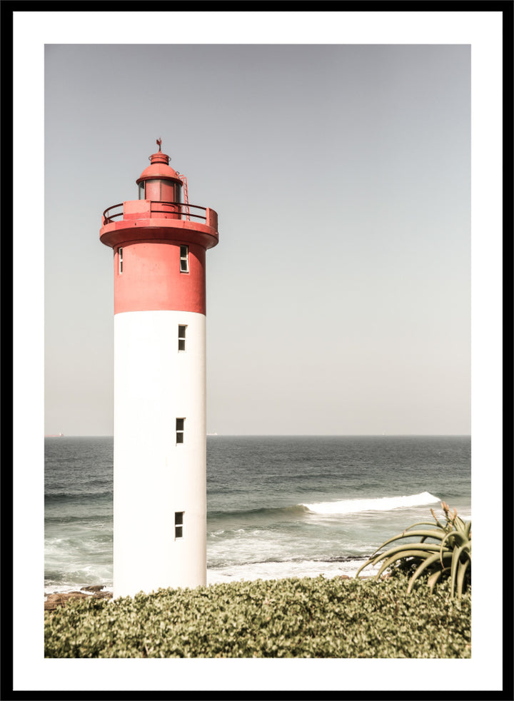 A framed photographic art print of a red and white lighthouse standing on a grassy shore with the ocean and sky in the background. Beacon by the Sea by Shot by Clint coastal photography print featuring a red-and-white lighthouse, ocean waves, pale sky, and coastal greenery.