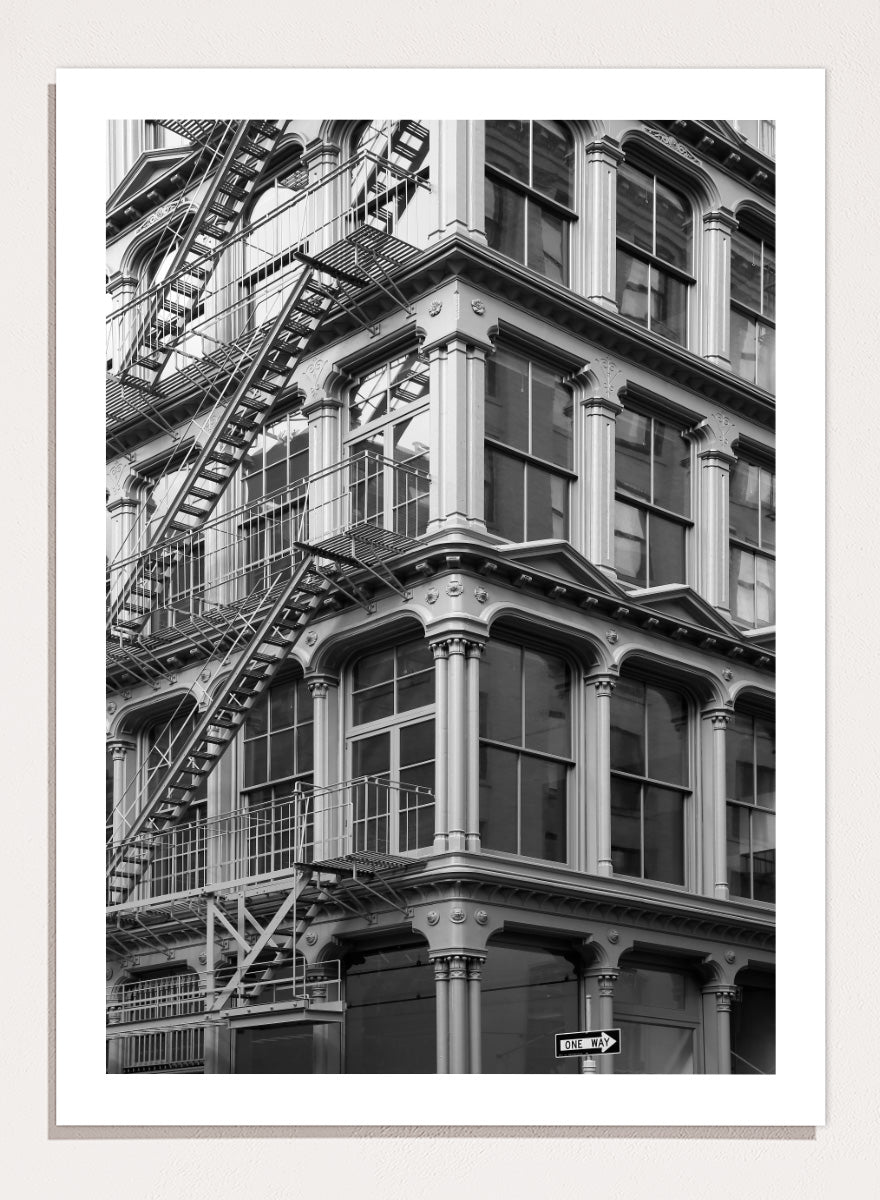 Black and white architectural photograph of a corner building facade with ornate windows and external fire escapes. Urban Geometry by Shot by Clint black-and-white architectural photography print featuring fire escapes, tall windows, ornate columns, and layered city detail.