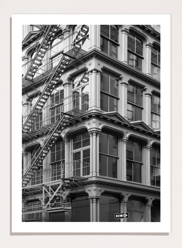 Black and white architectural photograph of a corner building facade with ornate windows and external fire escapes. Urban Geometry by Shot by Clint black-and-white architectural photography print featuring fire escapes, tall windows, ornate columns, and layered city detail.