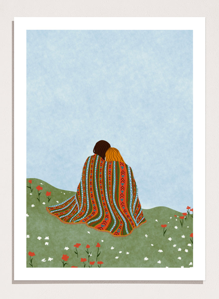 A framed art print featuring an illustration of two people sitting on a grassy hill, wrapped in a vibrant, patterned blanket under a light blue sky. Cozy Hillside Embrace by Ceyda Alasar art print featuring two people wrapped in a patterned blanket among green hills and wildflowers.