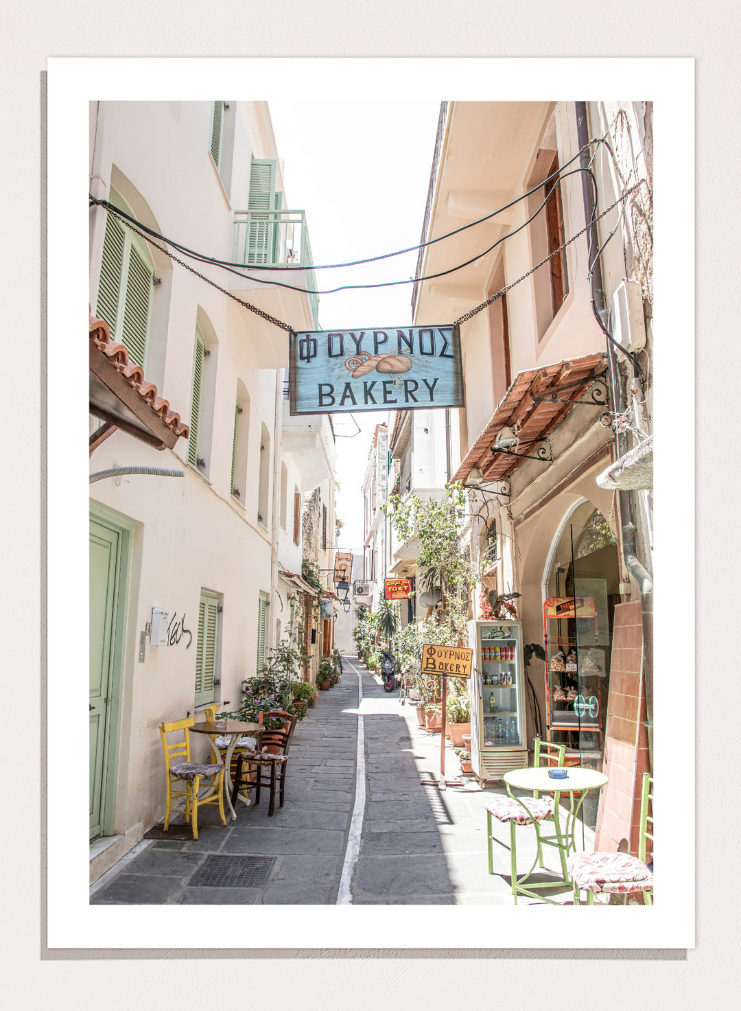  narrow street with a bakery sign in a European town. Charming Bakery Street by Henrike Schenk is a Greek street photography print featuring a Mediterranean bakery alley, pastel buildings, café seating, and travel wall art.