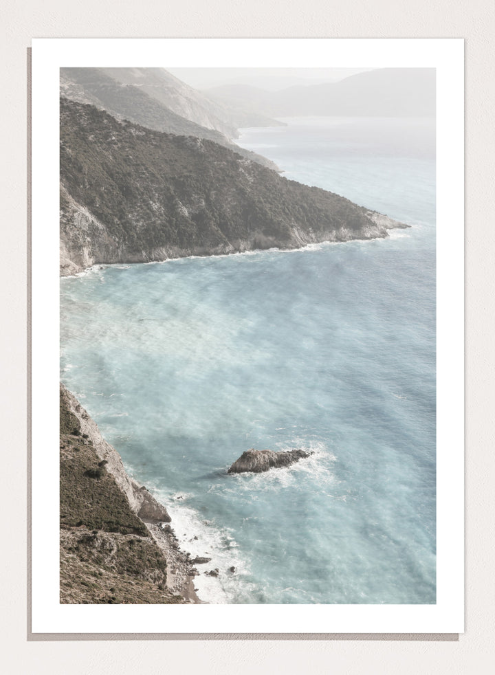 A framed photographic art print depicting a coastal landscape with a steep rocky cliff overlooking a calm, pale blue sea with a small rock formation in the foreground. Coastal Serenity by Shot by Clint coastal photography print featuring pale blue water, rugged cliffs, rocky shores, and a soft hazy horizon.