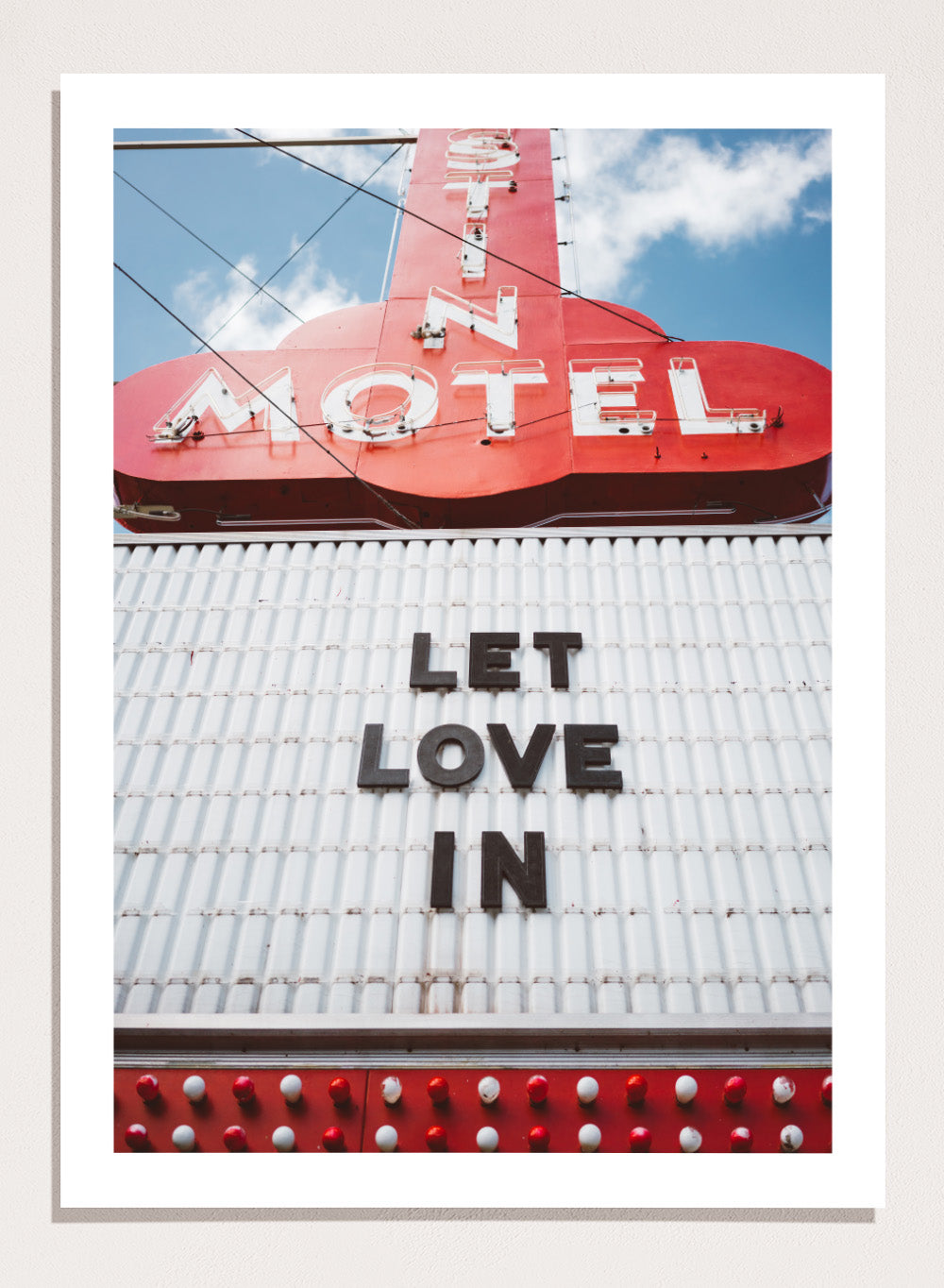 Vintage motel sign with 'Let Love In' message against a blue sky. Let Love In by Bethany Young features a vintage motel sign with red neon lettering against blue sky. Retro Americana photography wall art print.