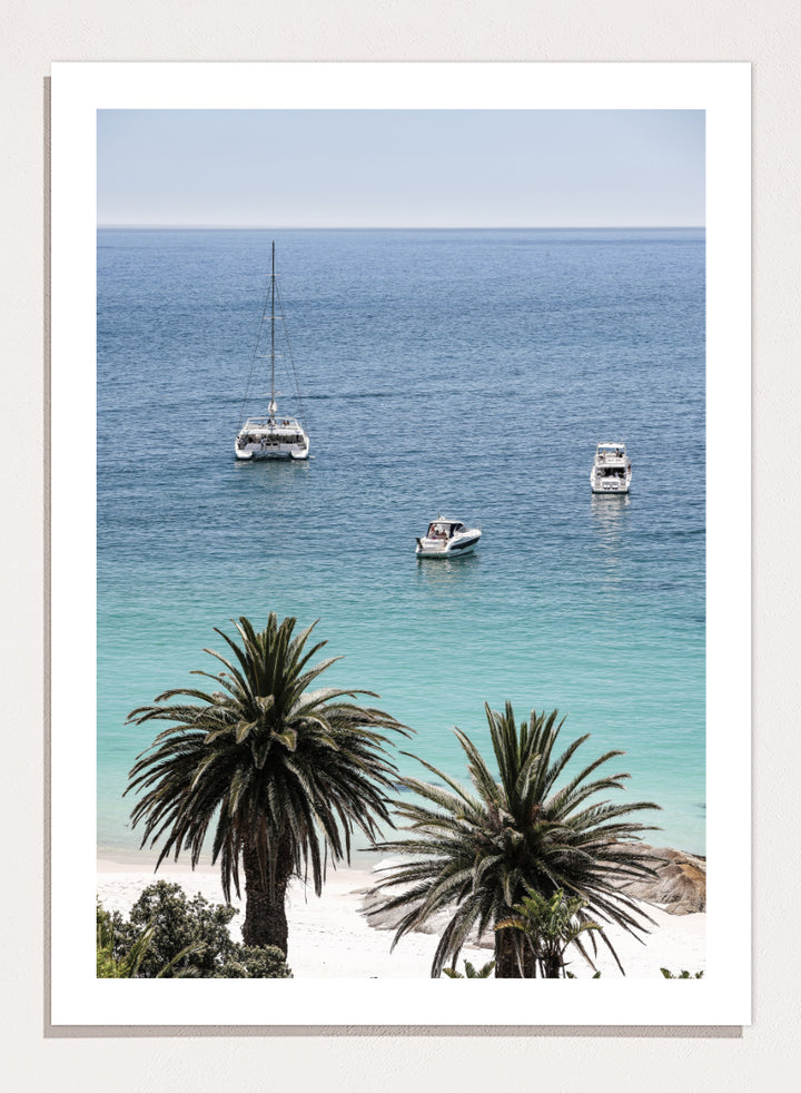 A framed coastal photograph showing a calm blue ocean with three boats, turquoise water near a white sandy shore, and two palm trees in the foreground. Tropical Serenity by Shot by Clint coastal photography print featuring turquoise water, sailboats, palm trees, and a bright tropical shoreline.