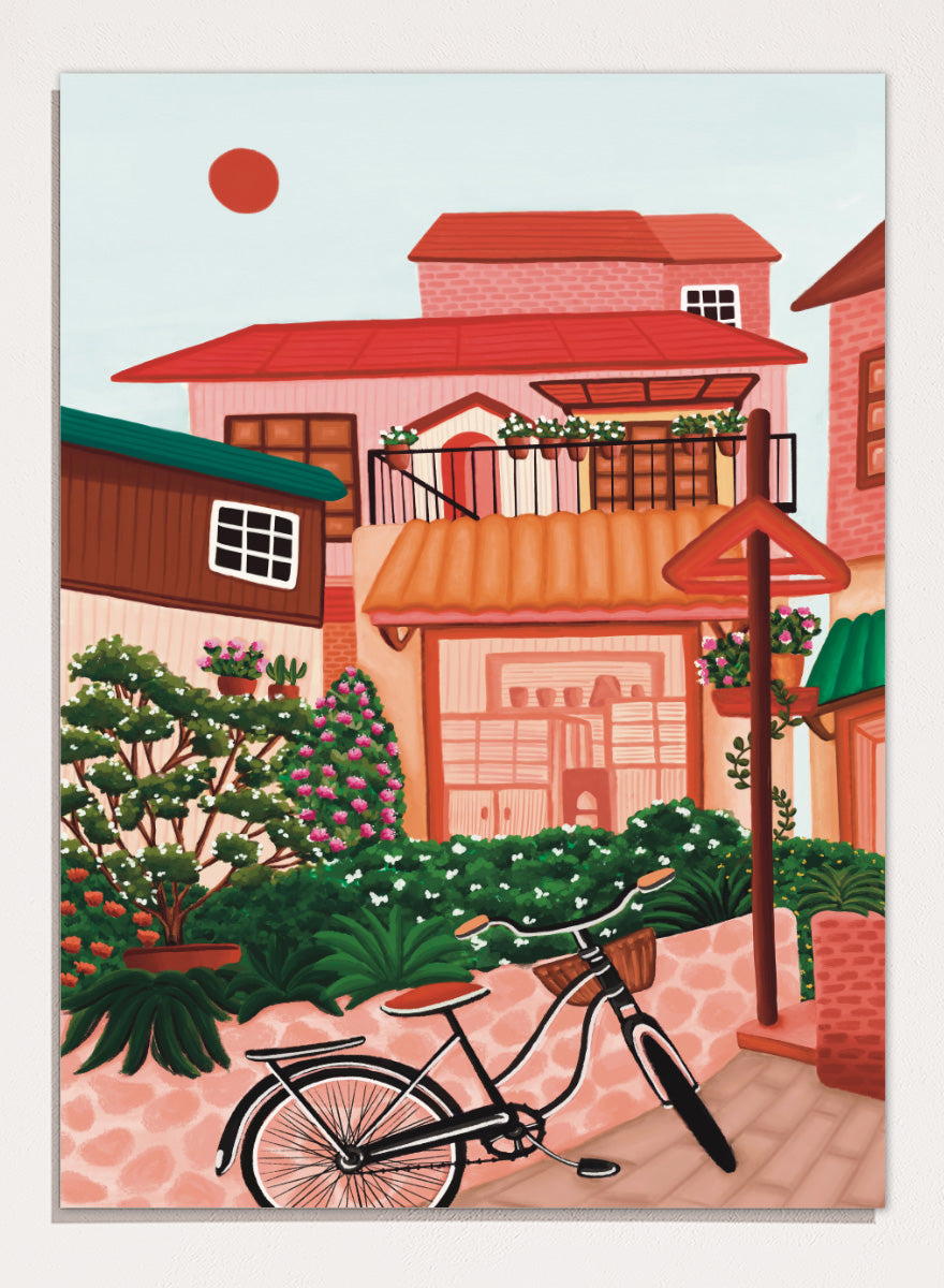 A colorful illustrated street scene with pink and peach buildings, green plants, flowers, a bicycle in the foreground, and a pale sky with a red sun. Shop Charming Village Street by Nidhi Kachhadiya, a colorful village wall art print featuring a bicycle, flower-filled homes, terracotta roofs, and warm pink and green tones.