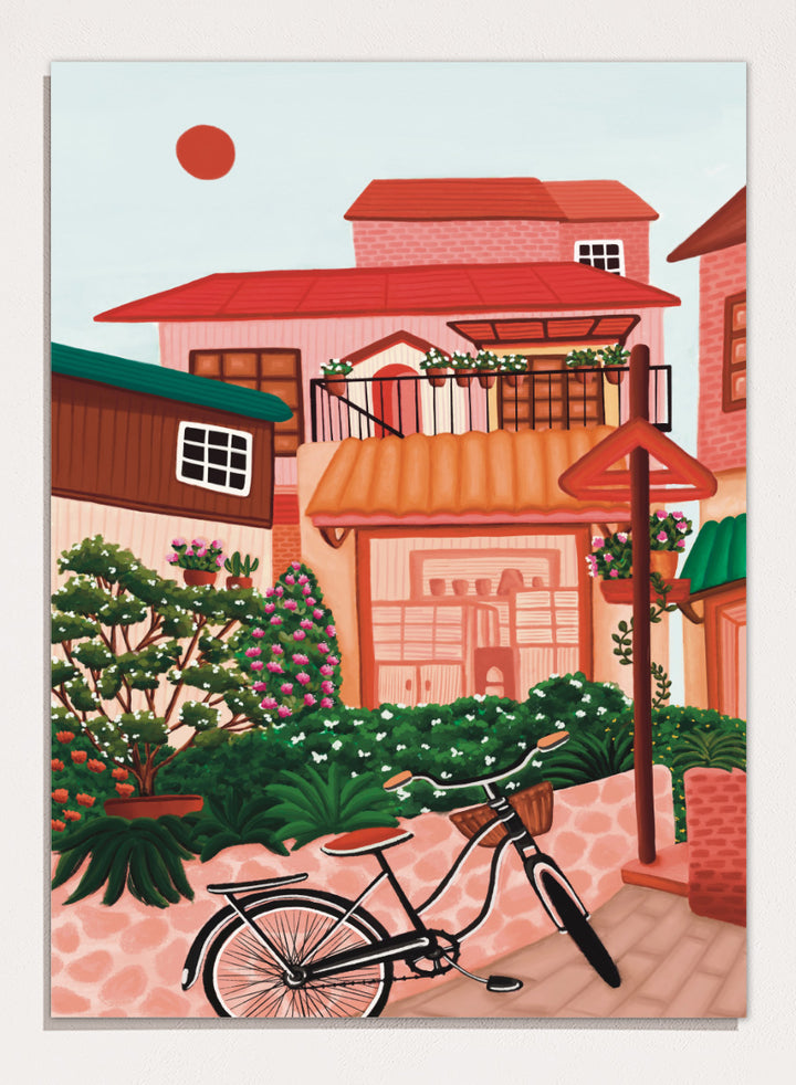 A colorful illustrated street scene with pink and peach buildings, green plants, flowers, a bicycle in the foreground, and a pale sky with a red sun. Shop Charming Village Street by Nidhi Kachhadiya, a colorful village wall art print featuring a bicycle, flower-filled homes, terracotta roofs, and warm pink and green tones.