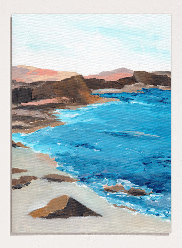 A framed landscape painting depicting a coastal scene with bright blue ocean water, sandy shore, dark rocky cliffs, and distant hills under a pale sky. Shop Rocky Coastal Waters by Nikita Jariwala, a coastal wall art print poster with blue ocean water, rocky cliffs, and a peaceful seaside view.