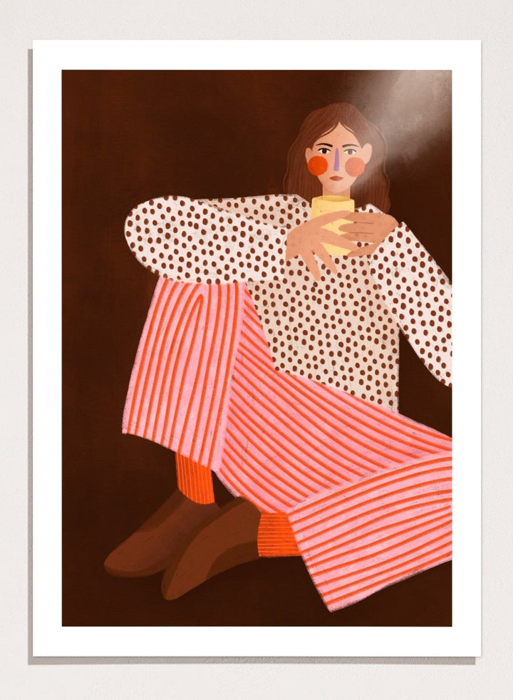 A framed art print featuring a stylized illustration of a woman in a white blouse with brown polka dots and pink-and-white striped trousers, holding a yellow cup against a dark brown background. Cozy Coffee Moment by Bea Müller premium framed fine art print featuring a woman curled up with a warm mug in bold pink, red, and brown tones.