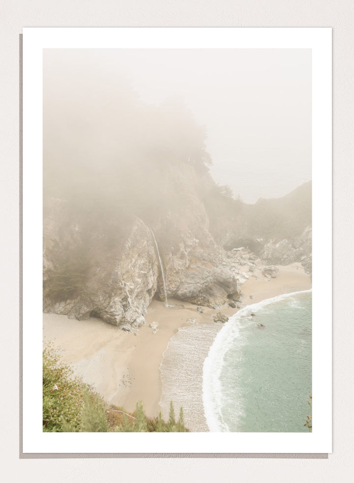 A framed photographic print of a misty coastal beach scene with a small waterfall, sandy shore, and pale blue-green ocean waves. Misty Coastal Haven by Shot by Clint coastal photography print featuring a secluded cove, rocky cliffs, pale turquoise water, and a soft veil of mist.