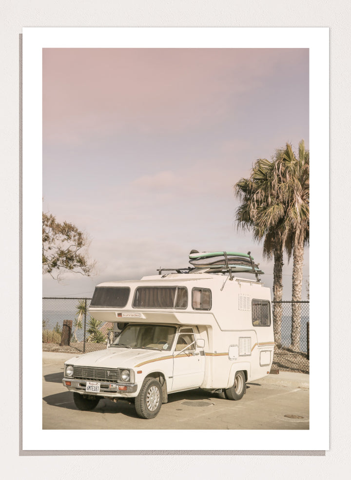 A vintage white camper truck parked outdoors near palm trees and a fence, with a surfboard strapped to its roof rack. Vintage Camper Parked by Shot by Clint travel photography print featuring a retro camper, surfboards, palm trees, and sun-faded coastal tones.