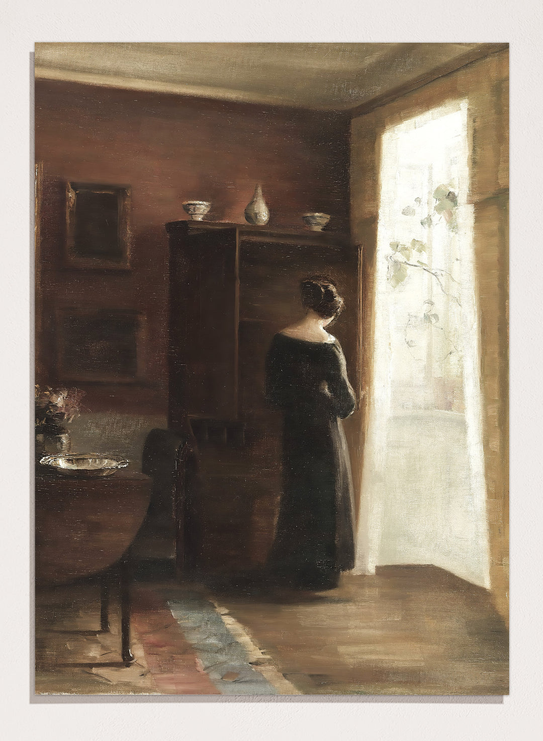 Painting of a woman standing in a sunlit room with a large mirror. Interior with a Young Girl Standing by the Window by Carl Holsøe is a vintage Danish interior art print featuring Scandinavian interiors, muted tones, and a woman by a window.