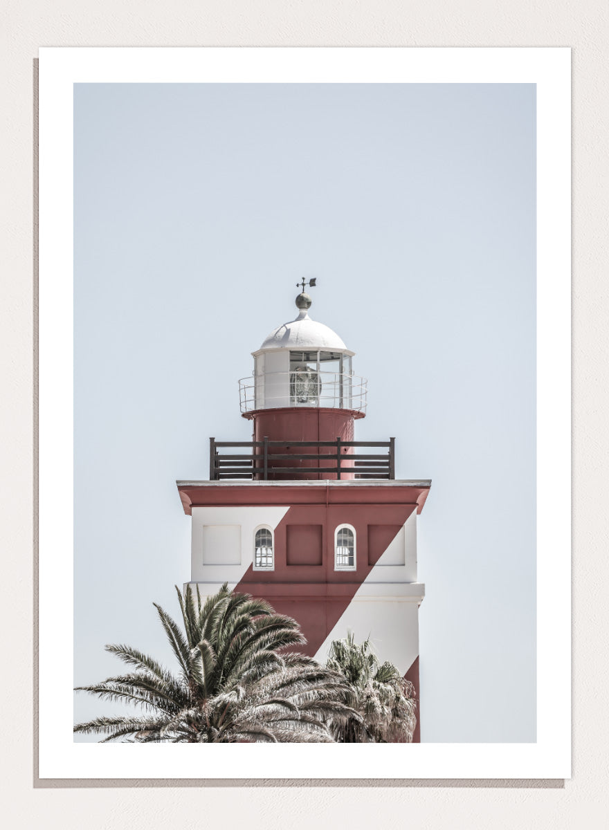 A framed art print featuring a red and white lighthouse with palm trees at the bottom, set against a pale sky background. Beacon of Solitude by Shot by Clint lighthouse photography print featuring red-and-white architecture, palm trees, arched windows, and a pale coastal sky.