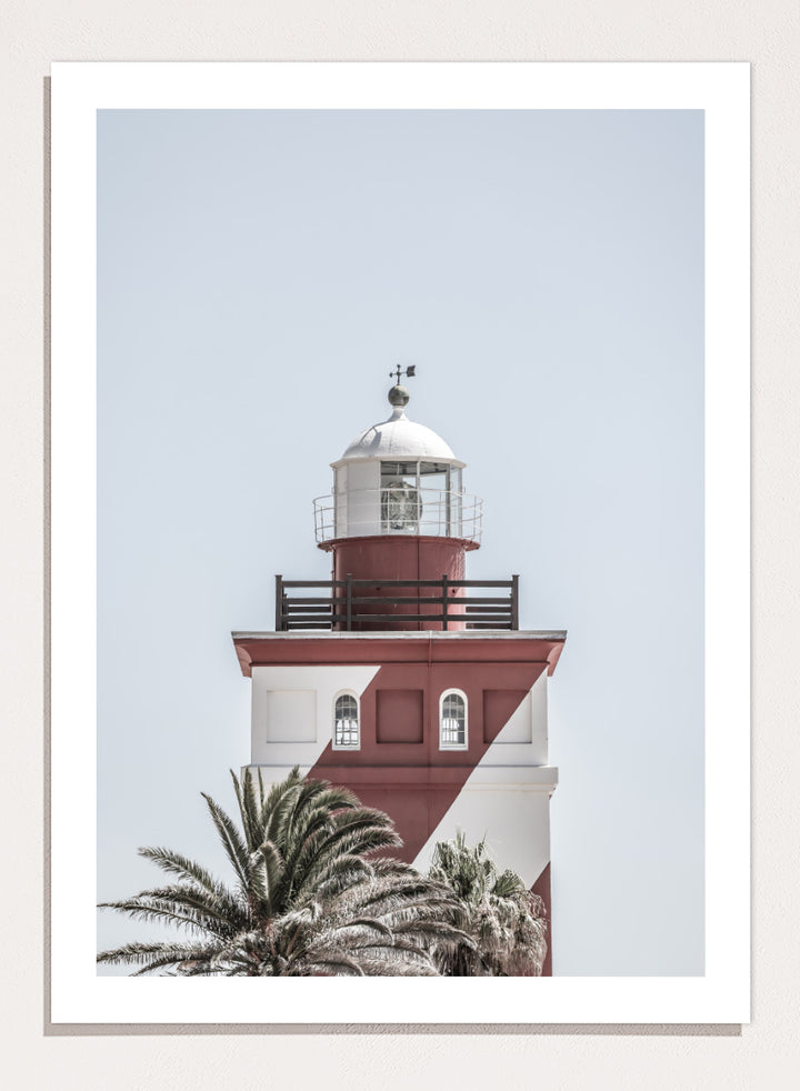 A framed art print featuring a red and white lighthouse with palm trees at the bottom, set against a pale sky background. Beacon of Solitude by Shot by Clint lighthouse photography print featuring red-and-white architecture, palm trees, arched windows, and a pale coastal sky.