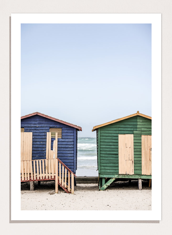 A framed photographic print of two wooden beach huts on a sandy beach under a pale blue sky, with one hut in dark blue and the other in green, both with light wood shutters and doors. Coastal Huts Harmony by Shot by Clint coastal photography print featuring blue and green beach huts, sandy ground, weathered wood, and ocean views.