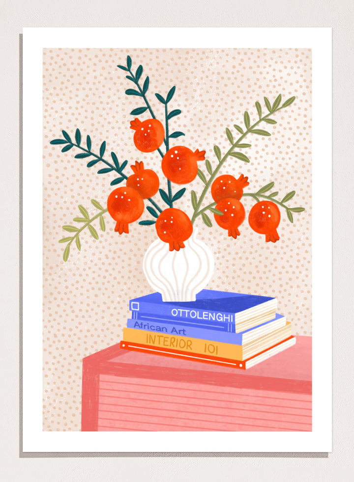 A framed art print featuring a stylized illustration of pomegranate branches in a white vase resting on a stack of books, set against a light beige dotted background. Cozy Tabletop Pomegranates by Bea Müller still life art print featuring red fruit branches, a sculptural vase, design books, and colorful interior styling.
