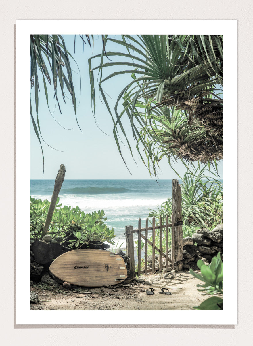 A framed beach scene art print featuring a wooden surfboard leaning against a rustic gate, with palm leaves framing a view of the ocean under a light blue sky. Coastal Serenity by Shot by Clint coastal art print featuring a surfboard, tropical plants, rustic gate, sandy path, and ocean waves.