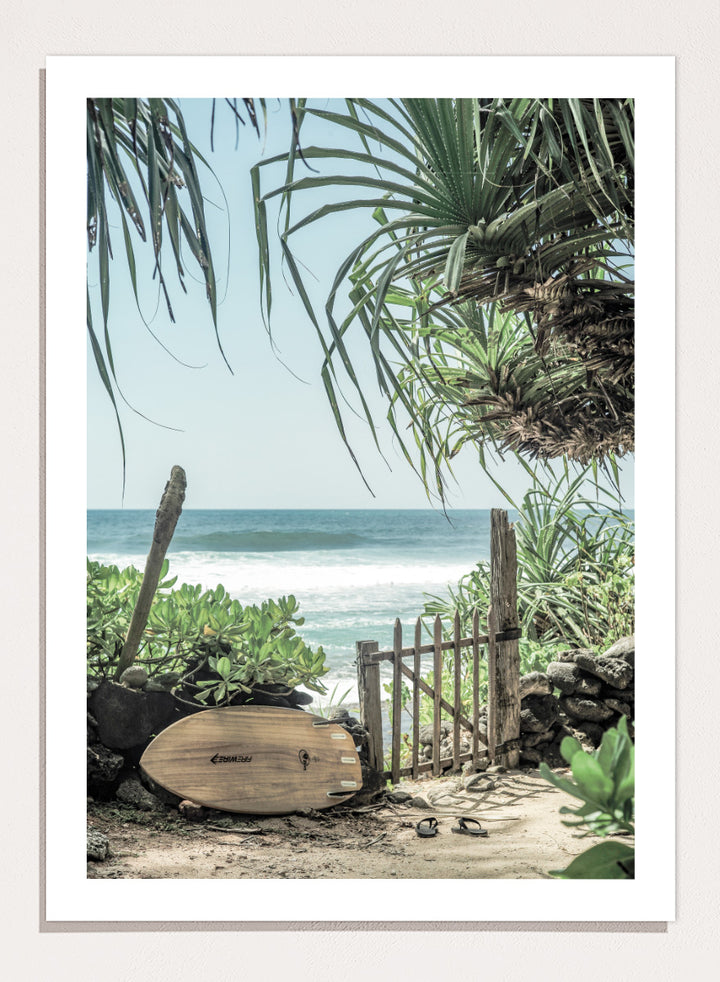 A framed beach scene art print featuring a wooden surfboard leaning against a rustic gate, with palm leaves framing a view of the ocean under a light blue sky. Coastal Serenity by Shot by Clint coastal art print featuring a surfboard, tropical plants, rustic gate, sandy path, and ocean waves.