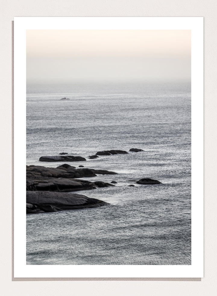 A framed black and white coastal landscape photograph featuring a calm sea, distant rocks, and a pale sky. Silver Sands by Shot by Clint coastal photography print featuring silver-blue water, dark shoreline rocks, and a soft misty horizon.
