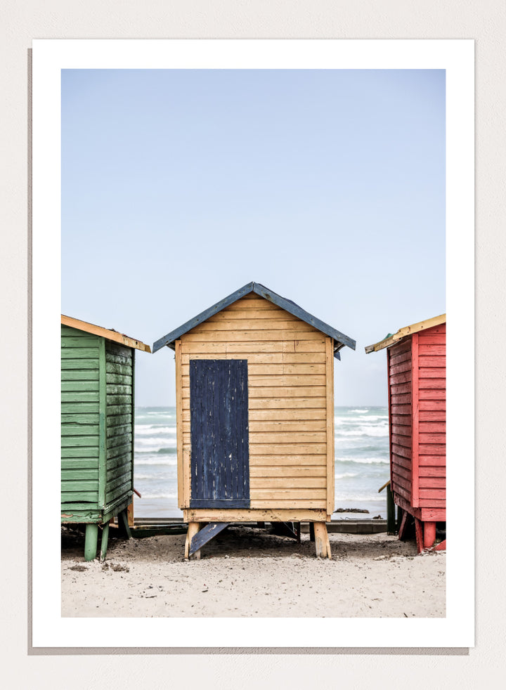 A framed photographic print of three colorful wooden beach huts on a sandy beach, with the central hut in natural wood and a dark blue door, flanked by green and red huts under a pale blue sky. Colorful Beach Huts by Shot by Clint coastal photography print featuring green, natural wood, and red huts with weathered texture and ocean views.