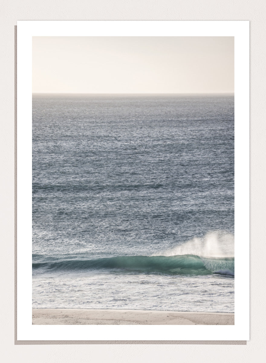 A framed photographic art print of an ocean scene, featuring a gentle wave breaking on a sandy beach under a pale sky. Tranquil Ocean Scene by Shot by Clint ocean photography print with a rolling wave, soft sea spray, and airy blue coastal tones.
