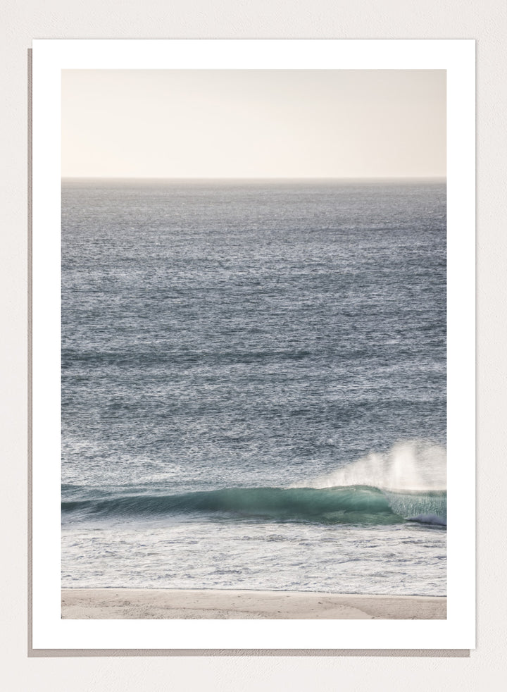 A framed photographic art print of an ocean scene, featuring a gentle wave breaking on a sandy beach under a pale sky. Tranquil Ocean Scene by Shot by Clint ocean photography print with a rolling wave, soft sea spray, and airy blue coastal tones.