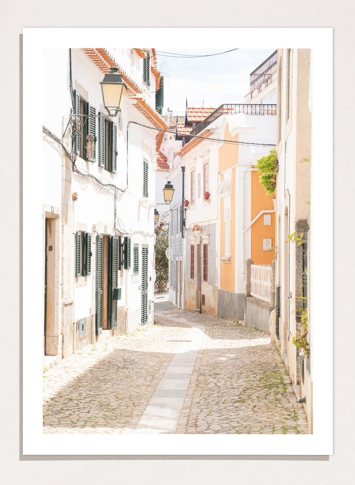 Cobbled street in a quaint European town with white buildings and a clear sky. Charming European Cobblestone Street by Henrike Schenk is a travel photography print featuring cobblestone streets, historic architecture, and European wall art.