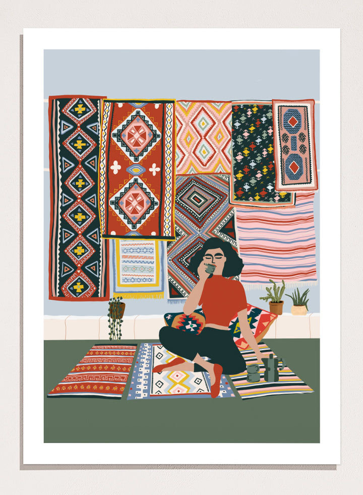 A framed art print featuring a stylized illustration of a woman sitting cross-legged on a patterned rug, surrounded by colorful geometric textiles hanging on a wall. Bohemian Coffee Morning by Ceyda Alasar interior art print featuring patterned rugs, floor cushions, potted plants, coffee, and colorful collected decor.